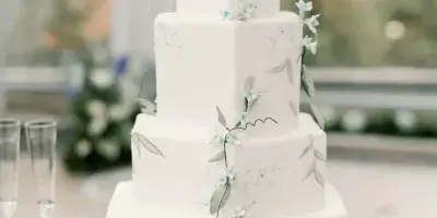 A four-tier white wedding cake decorated with hand-painted floral vines and fresh flowers, displayed on a table with champagne flutes.