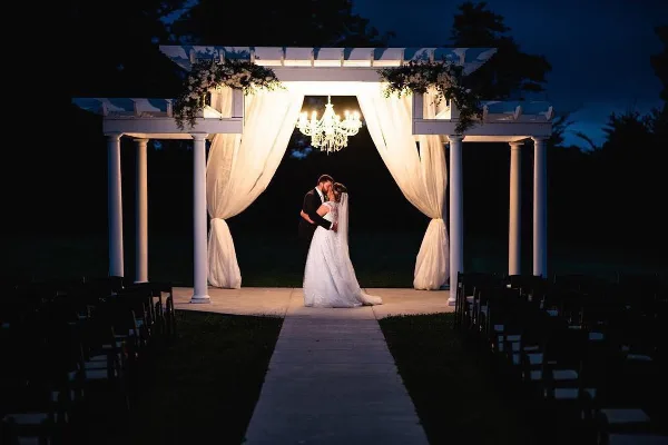 A couple standing under a white wedding arch with drapery and a chandelier at dusk, with rows of chairs set up for a ceremony.