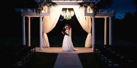 A couple standing under a white wedding arch with drapery and a chandelier at dusk, with rows of chairs set up for a ceremony.