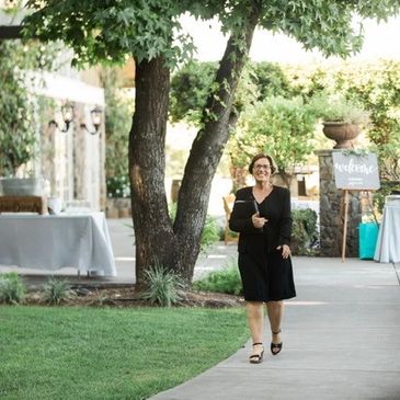 A woman in a black dress stands on a pathway in an outdoor venue, holding a book.