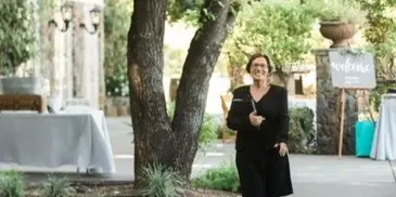 A woman in a black dress stands on a pathway in an outdoor venue, holding a book.