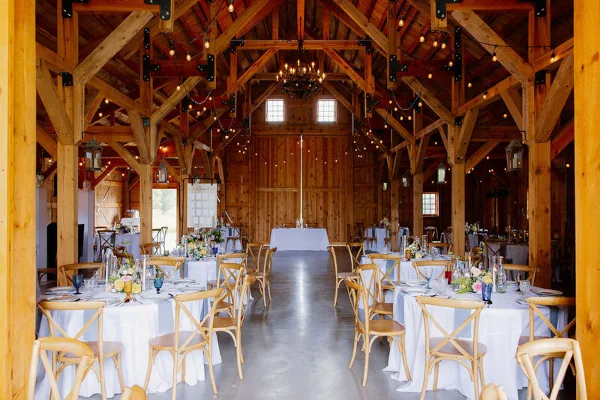 Interior of a wooden barn venue set up for a wedding reception with round tables and string lights.