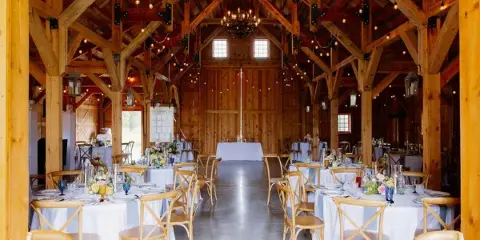 Interior of a wooden barn venue set up for a wedding reception with round tables and string lights.
