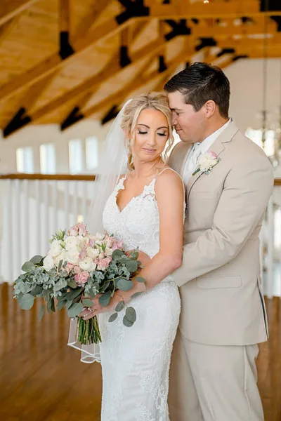 A bride in a white lace gown holding a bouquet of pink and white flowers stands with a groom in a beige suit inside a wooden-beamed venue.