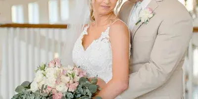 A bride in a white lace gown holding a bouquet of pink and white flowers stands with a groom in a beige suit inside a wooden-beamed venue.