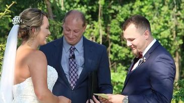 A bride and groom exchanging vows outdoors with an officiant standing between them.