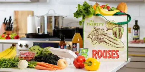 A Rouses Markets reusable grocery bag filled with fresh produce, including bananas, lettuce, and bread, sitting on a kitchen counter surrounded by other groceries like artichokes, carrots, and peppers.