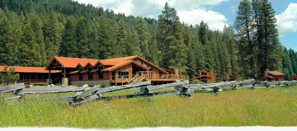 A rustic wooden lodge and cottages with red roofs situated in a grassy meadow, backed by a dense evergreen forest and mountains.