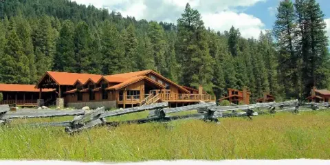 A rustic wooden lodge and cottages with red roofs situated in a grassy meadow, backed by a dense evergreen forest and mountains.