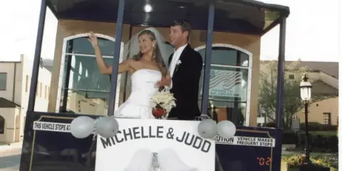 A bride and groom standing on a decorated trolley with a sign reading 'MICHELLE & JUDD'.