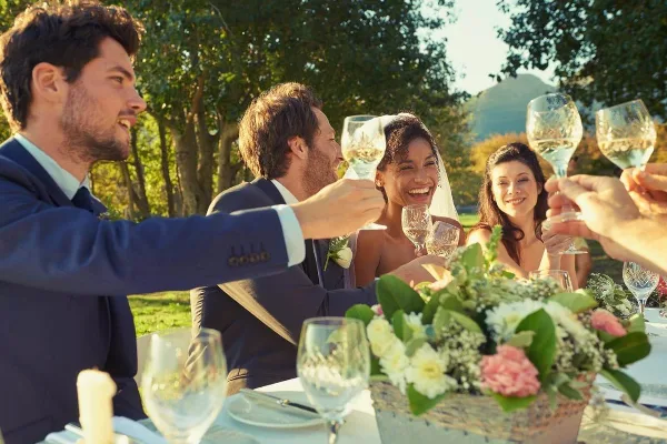 Wedding guests seated at a table with white linens and floral centerpieces toasting with wine glasses outdoors.