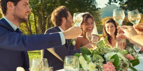 Wedding guests seated at a table with white linens and floral centerpieces toasting with wine glasses outdoors.
