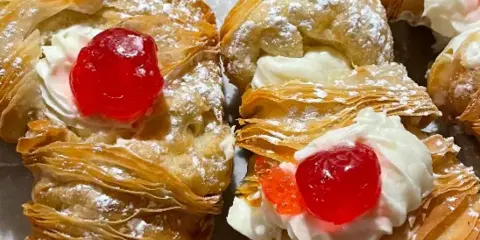 Close-up of a flaky pastry topped with whipped cream and a red cherry.
