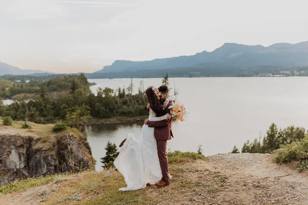 A bride and groom embracing on a cliff overlooking a lake and mountains.