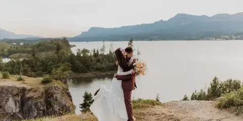 A bride and groom embracing on a cliff overlooking a lake and mountains.