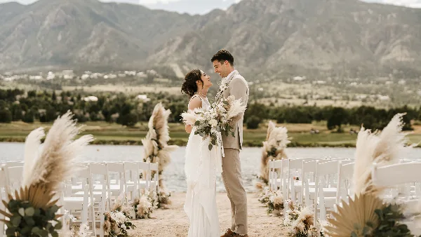 A bride and groom standing at a wedding ceremony on a beach with white chairs and mountain views.