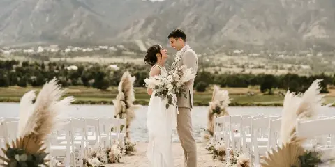 A bride and groom standing at a wedding ceremony on a beach with white chairs and mountain views.