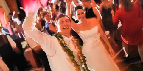 A bride and groom dancing at a wedding reception with a crowd of guests in the background.