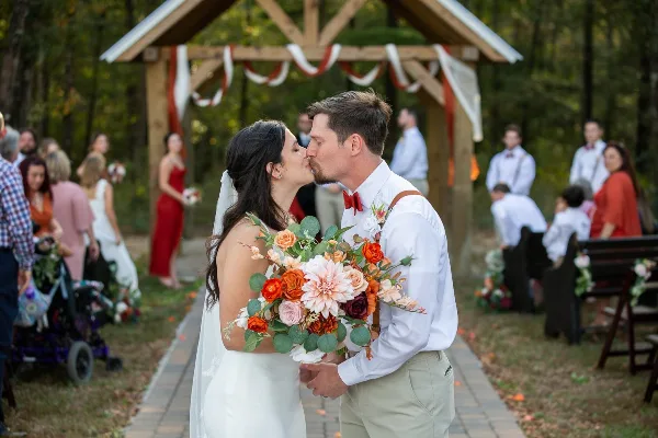 A bride and groom kissing during an outdoor wedding ceremony under a wooden arbor.