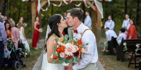 A bride and groom kissing during an outdoor wedding ceremony under a wooden arbor.