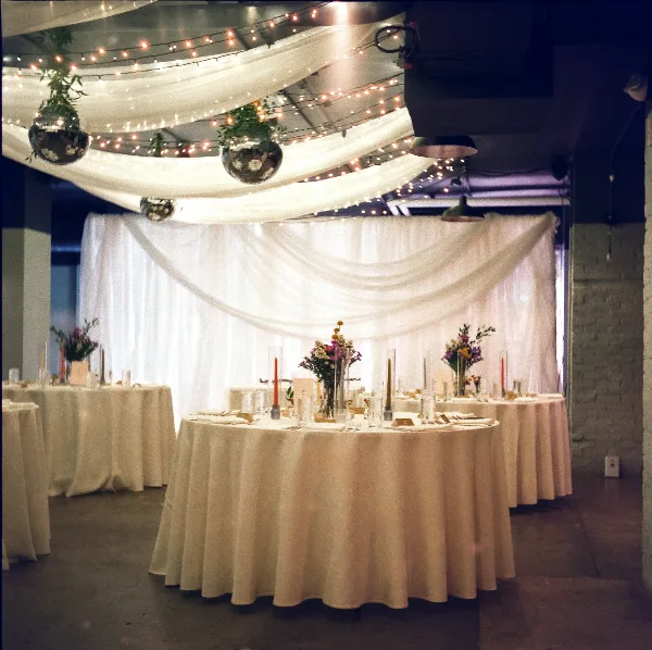 A wedding reception venue decorated with white ceiling drapery, string lights, and round tables with floral centerpieces.