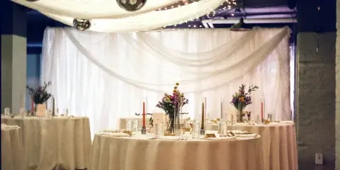 A wedding reception venue decorated with white ceiling drapery, string lights, and round tables with floral centerpieces.