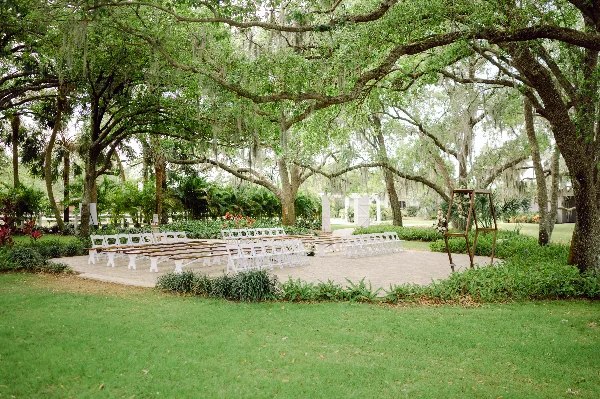 An outdoor wedding ceremony area with white chairs and a floral arch set up under large oak trees on a grassy lawn.