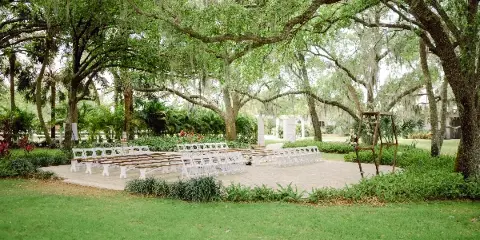 An outdoor wedding ceremony area with white chairs and a floral arch set up under large oak trees on a grassy lawn.