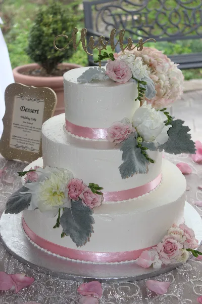 A three-tiered white wedding cake decorated with pink and white flowers, sage leaves, and a gold 'Mr & Mrs' topper.