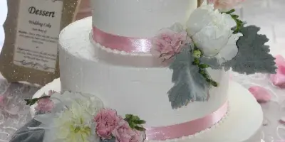 A three-tiered white wedding cake decorated with pink and white flowers, sage leaves, and a gold 'Mr & Mrs' topper.