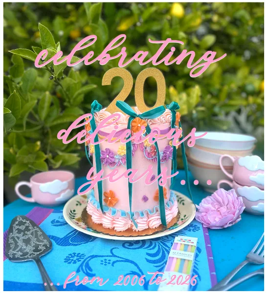 A pink custom cake decorated with ribbons and flowers, topped with a gold number 20 topper, sitting on a table with vintage teacups.