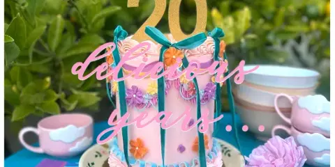 A pink custom cake decorated with ribbons and flowers, topped with a gold number 20 topper, sitting on a table with vintage teacups.