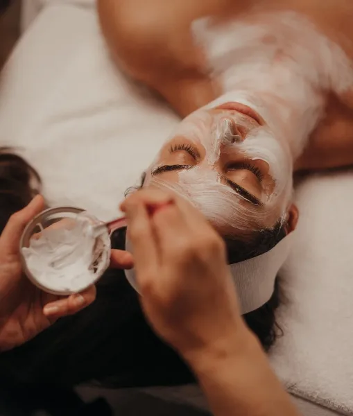 Aesthetician applying a white facial mask to a client's face during a spa treatment.