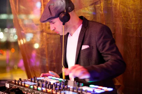 A DJ wearing a flat cap and headphones mixing music on a professional controller with illuminated buttons.