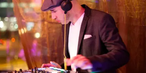 A DJ wearing a flat cap and headphones mixing music on a professional controller with illuminated buttons.
