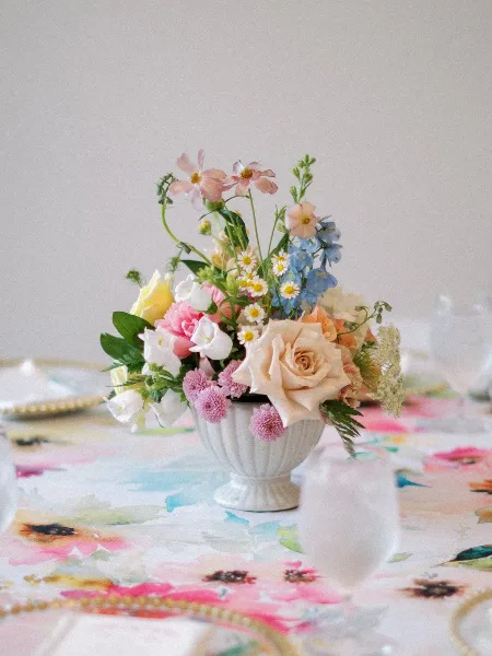 A floral wedding centerpiece featuring roses and daisies in a white urn on a floral tablecloth.