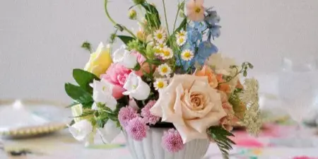 A floral wedding centerpiece featuring roses and daisies in a white urn on a floral tablecloth.
