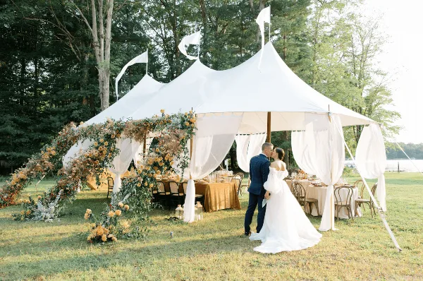 A white sailcloth tent with a peaked roof and flags set up outdoors for a wedding, featuring a floral arch and a couple kissing inside.