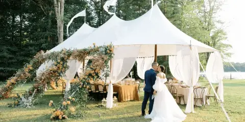 A white sailcloth tent with a peaked roof and flags set up outdoors for a wedding, featuring a floral arch and a couple kissing inside.