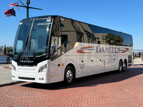 A white Prevost motor coach with 'DANIELS' branding parked on a brick surface near a waterfront.