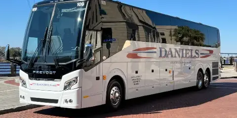 A white Prevost motor coach with 'DANIELS' branding parked on a brick surface near a waterfront.