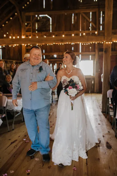 A bride in a white lace gown holding a bouquet walks through a rustic wooden barn interior with string lights and guests.