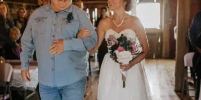 A bride in a white lace gown holding a bouquet walks through a rustic wooden barn interior with string lights and guests.