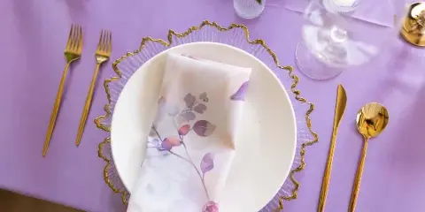 Lavender table setting with gold cutlery, white plates, and floral napkins on a lilac tablecloth