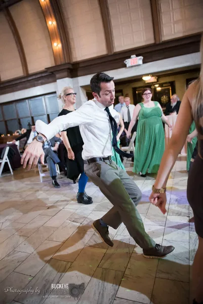 A wedding DJ in a white shirt and tie dancing on a ballroom dance floor with guests around him.