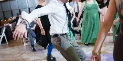 A wedding DJ in a white shirt and tie dancing on a ballroom dance floor with guests around him.