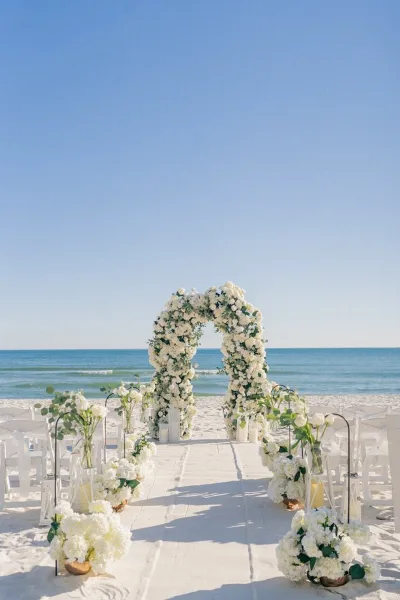 A white floral arch and aisle set up on a sandy beach for a wedding ceremony.