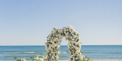 A white floral arch and aisle set up on a sandy beach for a wedding ceremony.
