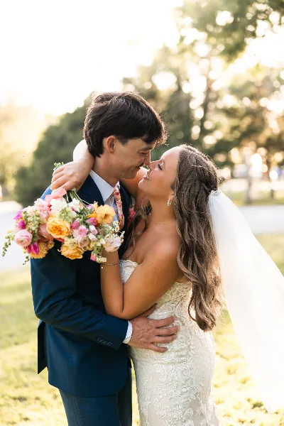 A bride and groom embracing and smiling at each other outdoors, with the bride holding a colorful bouquet.