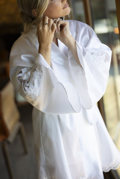 Bride in a white lace robe fastening an earring during wedding day preparations